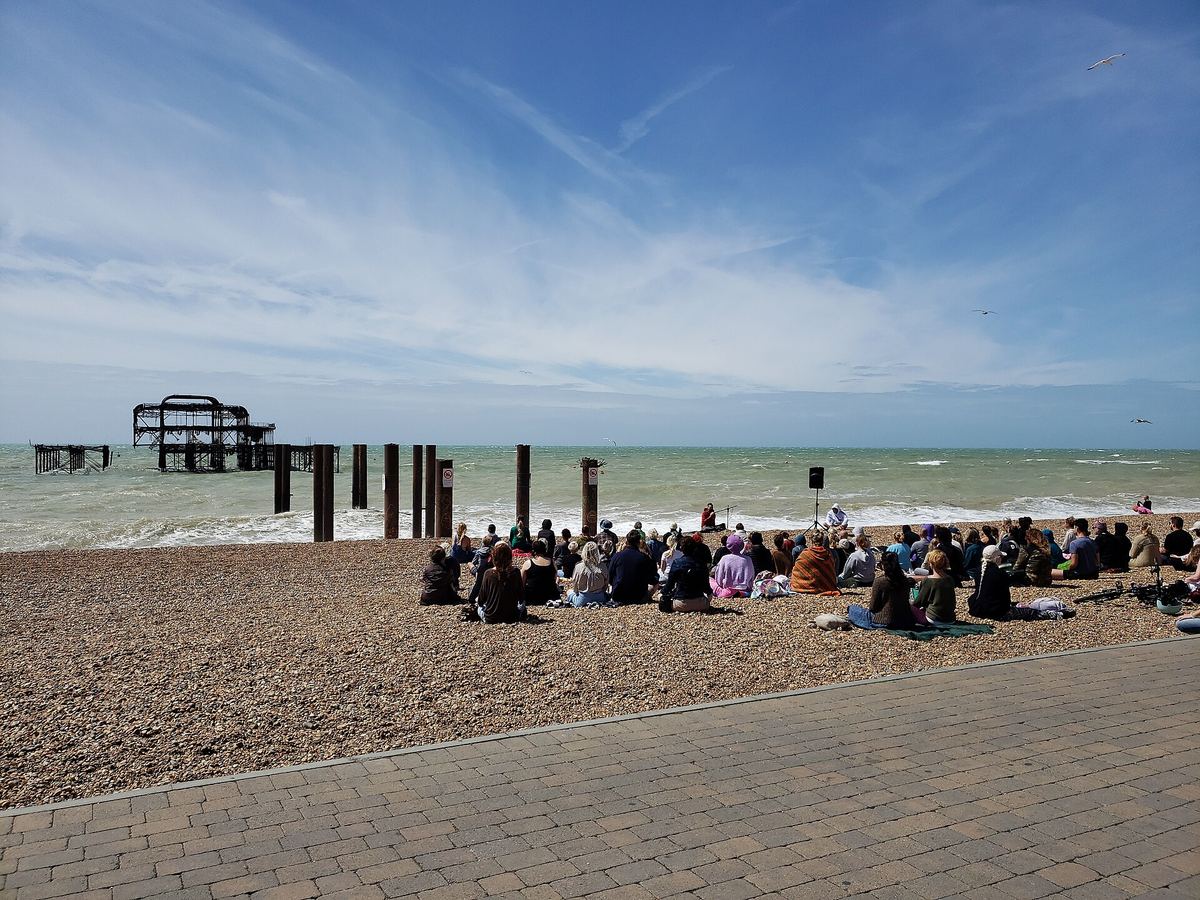 Eine Gruppe von Menschen sitzt meditierend am Kiesstrand vor einem alten Pier